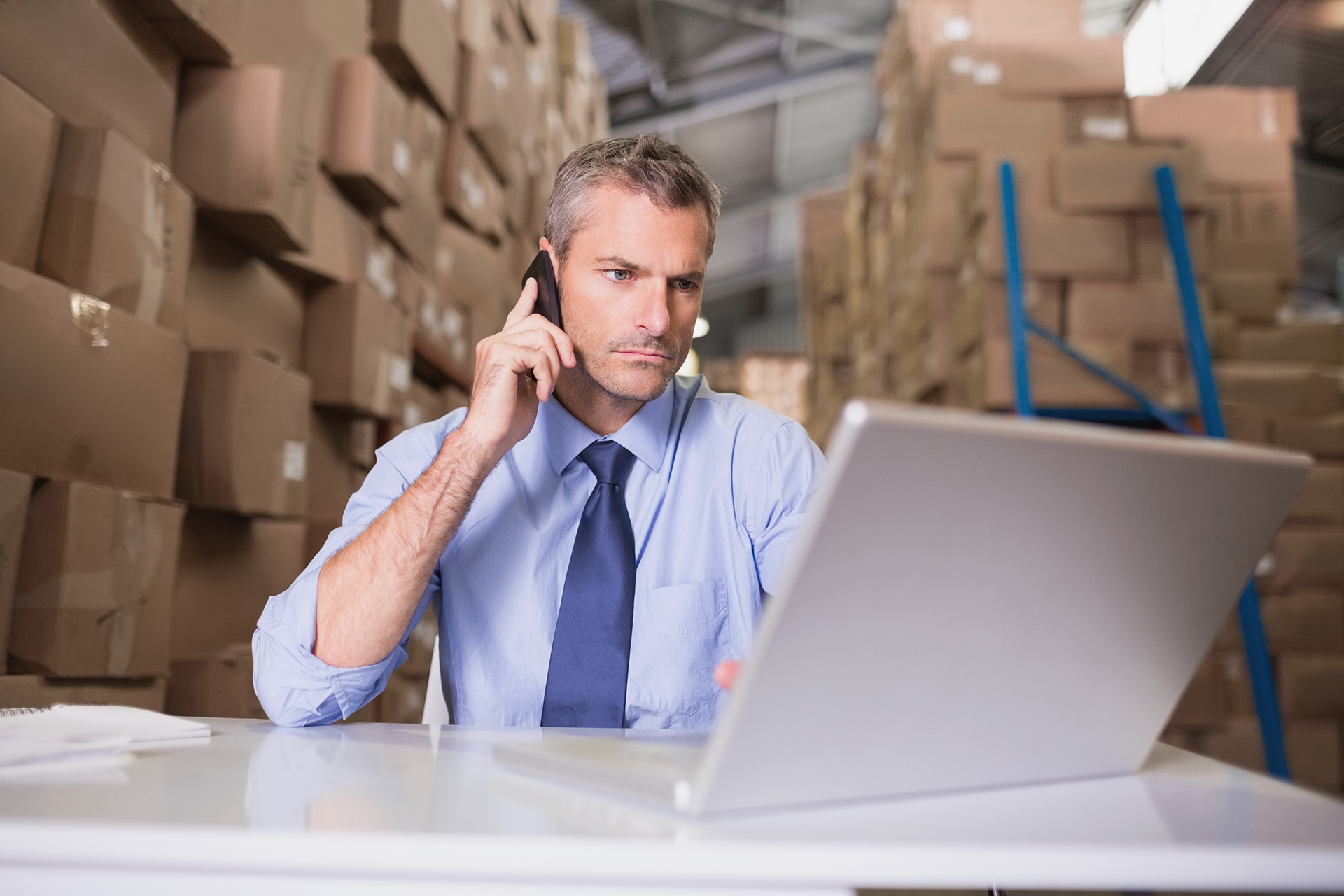 Warehouse manager using mobile phone and laptop at desk